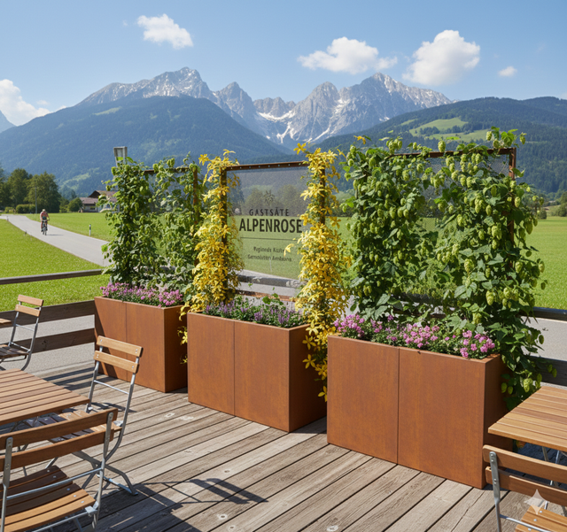 Foto einer Außenterrasse eines Gasthofes mit Holztischen und Blick auf ein Gebirgsmassiv. Als Begrenzung dienen drei große, rostbraune Cortenstahl-Pflanzkübel mit Rankgittern, an denen gelbe Kletterblumen und grüner Hopfen hochwachsen; das mittlere Gitter trägt ein Schild mit der Aufschrift „GASTSTÄTTE ALPENROSE