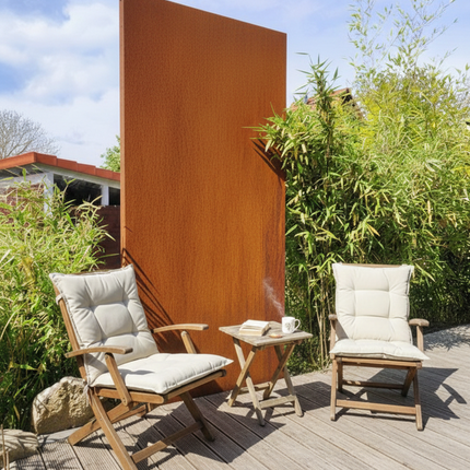 Eine sonnige Holzterrasse mit zwei klappbaren Gartenstühlen aus Holz und hellen Polstern, die vor einem hohen, rechteckigen Sichtschutzelement aus rostbraunem Cortenstahl stehen. Zwischen den Stühlen befindet sich ein kleiner Beistelltisch mit einem Buch und einer Tasse; im Hintergrund ist üppiges grünes Bambusgebüsch zu sehen.