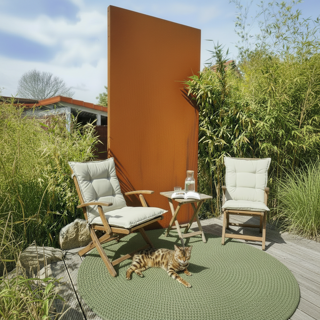 Eine sonnige Holzterrasse mit zwei klappbaren Gartenstühlen aus Holz und hellen Polstern, die vor einem hohen, rechteckigen Sichtschutzelement aus rostbraunem Cortenstahl stehen. Zwischen den Stühlen befindet sich ein kleiner Beistelltisch mit einem Buch und einer Tasse; im Hintergrund ist üppiges grünes Bambusgebüsch zu sehen.